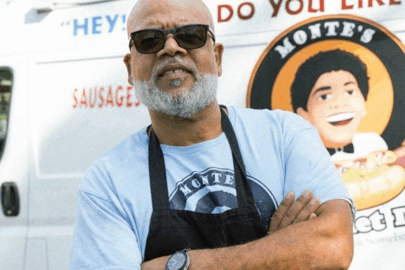 A man standing in front of a food truck wearing black sunglasses and apron.